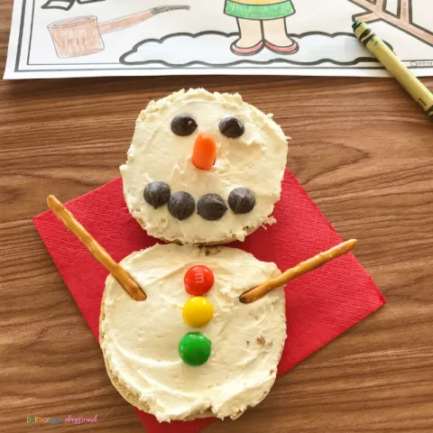 Picture of an open-faced bagel made to look like a snowman. White cream cheese with mini chocolate chips for eyes & mouth, carrot stick for a nose, three M&Ms for buttons, and 2 pretzel sticks for arms.